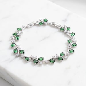 Peut inclure: Un bracelet de couleur argentée présentant un motif répétitif en forme de vigne. La pièce est ornée de multiples pierres ovales vert foncé et de petites pierres d'accent rondes et transparentes disposées en un motif délicat et symétrique.