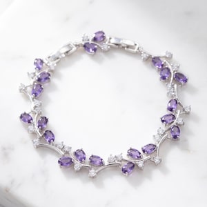 May include: A silver-toned bracelet featuring a repeating vine-like design. The metal links are set with marquise-cut purple amethyst stones and small, round, clear white stones arranged in a delicate, symmetrical pattern around the wrist band.