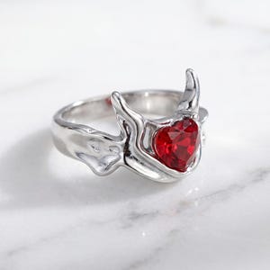 Peut inclure: Une bague argentée dotée d'un anneau en métal poli aux formes organiques, ornée de deux protubérances incurvées vers le haut rappelant des cornes. Une pierre précieuse rouge en forme de cœur facetté est sertie au centre de la monture.
