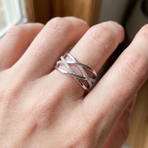 Peut inclure: Une bague argentée au design croisé multi-anneaux. La bague se compose de plusieurs brins de métal poli qui s'entrecroisent, dont l'un des brins centraux est incrusté de petites pierres claires et scintillantes sur sa surface visible.
