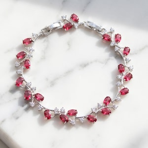 Peut inclure: Un bracelet argenté composé d'une série de pierres rouges ovales disposées en forme de vigne, entremêlées de petites pierres claires taillées en rond. Le bijou présente un fermoir métallique sécurisé et une finition métallique polie.