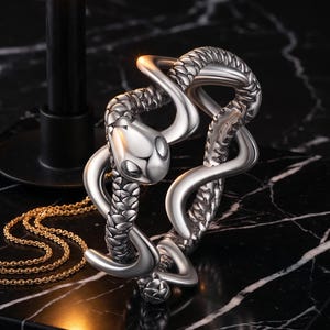 Peut inclure: Un bracelet en métal argenté en forme de serpent enroulé avec des textures d'écailles détaillées sur le corps et une tête lisse. Le bracelet est accompagné d'une fine chaîne de couleur dorée qui repose partiellement sous la forme métallique.