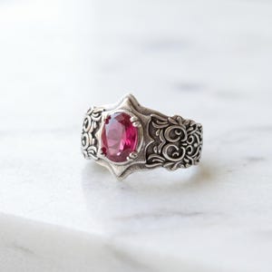 Può includere: Un anello in argento con una pietra preziosa di colore rosa intenso dal taglio ovale fissata con griffe. La fascia presenta intricati motivi a volute in rilievo che si estendono sulla superficie, circondando la pietra centrale in una cornice appuntita.