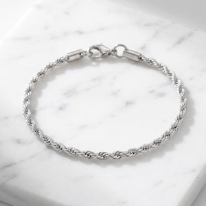 Peut inclure: Un bracelet chaîne torsadée de couleur argentée au design métallique entrelacé. Le bracelet est composé d'une série de maillons imbriqués créant un aspect texturé et spiralé, et est fixé par un fermoir mousqueton en métal poli.