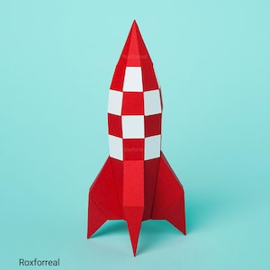 Op de afbeelding: Een papieren raketmodel met een rood en wit geblokt patroon op het hoofdgedeelte en effen rode vinnen aan de basis. De tekst Roxforreal staat op het lichaam en in de linkerbenedenhoek van de compositie afgedrukt.