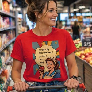 Peut inclure: T-shirt rouge avec le texte "An apple a day keeps anyone away if you throw it hard enough." Le t-shirt présente une illustration rétro d'une femme tenant une pomme. La personne pousse un chariot dans un supermarché.