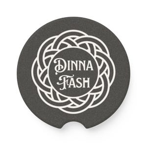 May include: A round, dark gray car coaster with a white Celtic knot design. The center of the coaster features the words "DINNA FASH" in a decorative font. The coaster has a small notch for easy removal from a car cup holder.