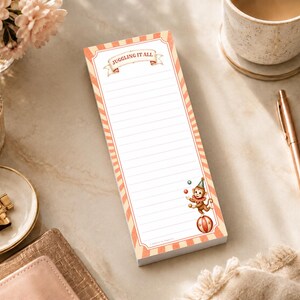 Puede incluir: Un bloc de notas rectangular con un centro rayado blanco y bordes a rayas naranjas y blancas. La parte superior presenta una pancarta con el texto JUGGLING IT ALL. La esquina inferior derecha muestra una pequeña ilustración de un mono.
