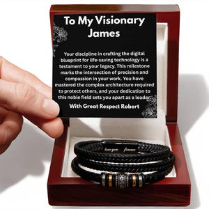 May include: A black braided leather bracelet with a metal clasp engraved with the words love you forever, featuring silver and gold-toned beads, presented in a dark wood box with a card reading To My Visionary James and a message from Robert.