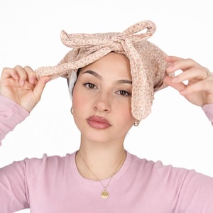 Op de afbeelding: Een lichtroze hoofddoek met een klein bloemenpatroon in tinten wit en bruin. De stof is aan de bovenkant vastgeknoopt met twee opvallende, langwerpige uiteinden die een strikvormige structuur vormen en de kruin van het hoofd bedekken.