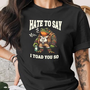 May include: Black t-shirt with a cartoon frog design. The frog is smoking and has tattoos, holding a beer can. The text reads "HATE TO SAY I TOAD YOU SO". The frog wears a vest with a skull on it.