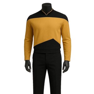 Puede incluir: Un uniforme de Star Trek amarillo mostaza y negro. La parte superior de manga larga presenta un canesú negro y un diseño de cheurón. El uniforme incluye pantalones negros y zapatos negros. El maniquí tiene guantes negros.