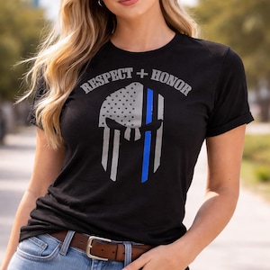 Puede incluir: Camiseta negra con un diseño de casco que incorpora la bandera estadounidense y una franja azul. El texto "RESPECT + HONOR" está impreso encima. La persona lleva vaqueros azules y un cinturón marrón.