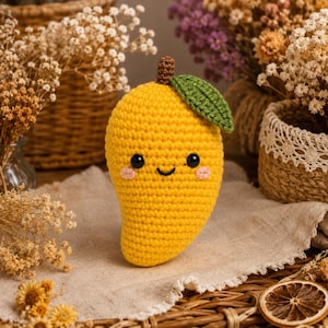May include: A yellow mango-shaped crochet plush toy featuring a brown stem, a single green leaf, two black safety eyes, and embroidered pink cheeks with a small black smile. The handmade textile fruit has a textured, stitched surface.