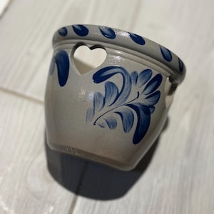 May include: A gray ceramic planter with blue floral designs and heart-shaped cutouts. The planter has a wide rim with blue leaf-shaped accents. The exterior features hand-painted blue floral patterns.