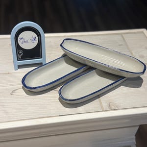 May include: Three ceramic serving dishes with a light gray interior and a dark blue rim. A small blue chalkboard sign with white text and a floral design is also present. The dishes are oval-shaped.