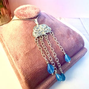 May include: Silver cloud pendant necklace with blue teardrop beads. The cloud pendant has detailed patterns and hangs from a fine chain. The necklace is displayed on a pink velvet surface.