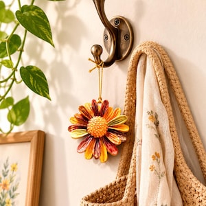 May include: A bronze-toned metal wall hook holds a hanging ornament shaped like a daisy flower with yellow, orange, and deep red petals. A beige woven tote bag with a floral patterned fabric lining hangs from the same hook on the wall.