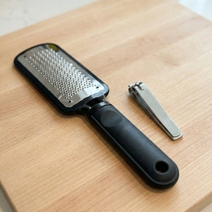Puede incluir: Una lima de pies negra con una superficie de acero inoxidable y un mango negro, junto a un cortaúñas plateado. Las herramientas están sobre una superficie de madera clara.