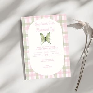 Könnte beinhalten: Eine rosa-grün karierte Einladungskarte mit dem Text "One Year Has Fluttered By" und einer Aquarell-Schmetterlingsillustration. Die Einladung enthält Details für eine erste Geburtstagsfeier, einschließlich Datum, Uhrzeit und Ort.