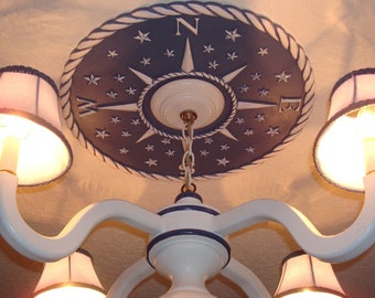 May include: A white ceiling medallion with a blue and white compass design and a rope border. The medallion is centered on a white ceiling and is surrounded by a white chandelier with five white lampshades.
