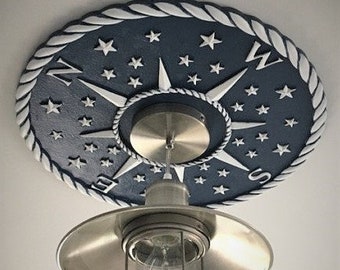 May include: A nautical-themed ceiling light fixture. The fixture features a navy blue oval base with a white rope-like border and star accents. A compass rose design is in the center. The light has a metal shade and a caged glass enclosure.