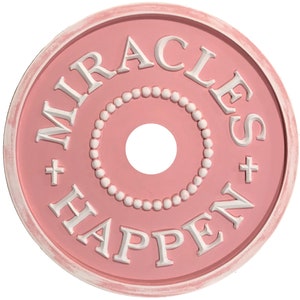 以下が含まれることがあります： 白の縁取りが付いたピンクの天井用メダルで、白文字で「MIRACLES + HAPPEN +」と書かれています。