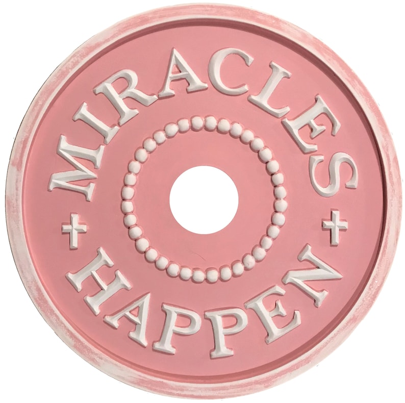 以下が含まれることがあります： 白の縁取りが付いたピンクの天井用メダルで、白文字で「MIRACLES + HAPPEN +」と書かれています。