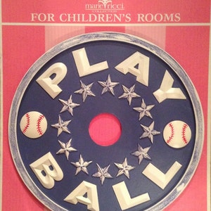 May include: A round, navy blue wall decoration with the words "PLAY BALL" in white, surrounded by silver stars and two baseballs. The decoration is set against a pink and white striped background, with the text "FOR CHILDREN'S ROOMS".