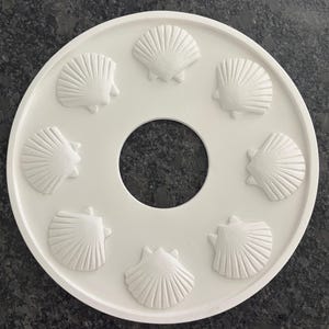 May include: A white, circular decorative tray with a central hole and eight raised seashell designs. The tray is made of a solid, opaque material and is set against a dark, speckled surface. The seashells are evenly spaced around the central hole.