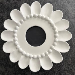 May include: A circular white frame shaped like a flower with rounded petals. The center features a smooth, circular opening surrounded by a ring of small, raised spherical beads. The overall design is symmetrical, matte white, and flat in profile.