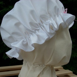 May include: A white cotton bonnet with a ruffled edge and a small pink bow. The bonnet is worn on a white fabric mannequin head.