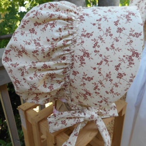 K&ouml;nnte beinhalten: Ein wei&szlig;er H&auml;ubchen mit rotem Blumenmuster. Das H&auml;ubchen ist aus Stoff und hat einen gerafften R&uuml;schenrand.