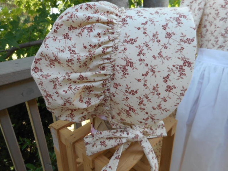 K&ouml;nnte beinhalten: Ein wei&szlig;er H&auml;ubchen mit rotem Blumenmuster. Das H&auml;ubchen ist aus Stoff und hat einen gerafften R&uuml;schenrand.