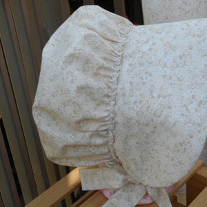 May include: A white bonnet with a floral pattern. The bonnet has a gathered ruffle around the crown and ties under the chin.