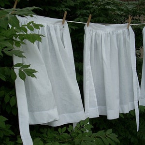 May include: Four white cotton aprons hanging on a clothesline outdoors. The aprons have a gathered ruffle at the top and are tied at the waist with long ties.