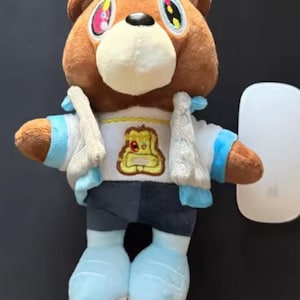 Puede incluir: Un peluche de oso pardo, vestido con una chaqueta blanca con detalles azules, una camisa blanca con un gráfico, pantalones cortos azul oscuro y botas azul claro. El oso tiene ojos coloridos y una nariz negra.