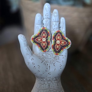 Könnte beinhalten: Ein Paar dreieckige Perlenohrringe mit einem zentralen, blumenartigen Motiv in Rot-, Orange- und Gelbtönen, umrandet von einem dunkelbraunen Rahmen und einer Außenkante aus kleinen weißen, gelben und orangefarbenen Perlen an silbernen Haken.