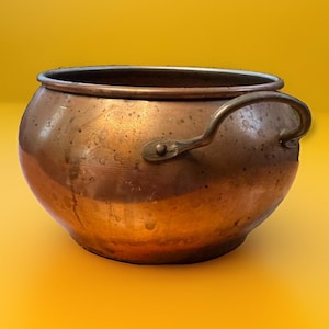 May include: A round copper pot with a wide opening and a single curved metal handle attached to the side with a rivet. The exterior surface of the pot shows a weathered, metallic finish with visible variations in color and texture.