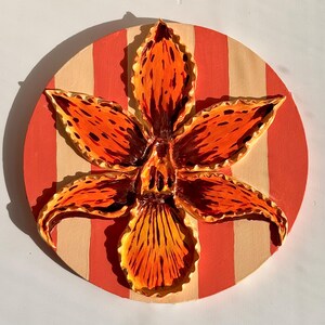 May include: A vibrant, orange and brown flower sculpture with detailed petals, set against a circular background of alternating red and beige stripes. The flower's intricate design and color palette create a striking visual effect, perfect for wall decor.