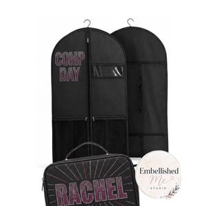 Op de afbeelding: Twee zwarte kledinghoezen en een zwarte rechthoekige accessoiretas. De linker kledinghoes heeft de tekst COMP DAY in roze strass-steentjes. De tas heeft de naam RACHEL in roze strass-steentjes met uitstralende lijnen.