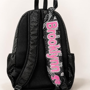 May include: A black glittery backpack with adjustable shoulder straps and a black bandana tied to one strap. The bandana features a white paisley pattern and the name Brooklynn in large, bright pink glittery letters with a silver border.