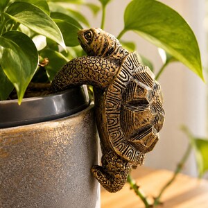 May include: A bronze-coloured turtle figurine with a textured shell and skin, designed to hook onto the rim of a plant pot. The turtle is positioned with its front legs over the edge of a grey, speckled ceramic pot containing a green leafy plant.