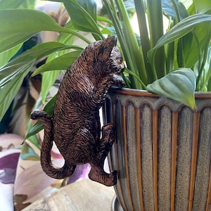 May include: A bronze cat ornament clinging to a plant pot. The cat has detailed fur texture and is positioned as if climbing. The plant pot is a muted green-brown with vertical grooves, and lush green plants are visible.