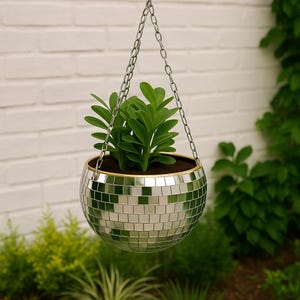May include: A hanging planter with a disco ball design, featuring a mosaic of silver tiles. The planter holds green plants and is suspended by a silver chain. The rim has a gold trim. The background includes a white brick wall and green foliage.