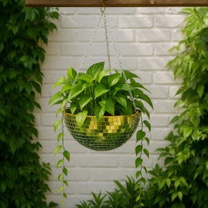 May include: A hanging planter with a gold mosaic finish, filled with vibrant green plants. The planter is suspended by a silver chain against a white brick wall and lush greenery. The trailing vines add a decorative touch.