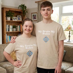 May include: Two people wearing tan t-shirts with the text "My Brain My Rules" and a blue and gray geometric design. They are standing in a living room with a couch and bookshelf.