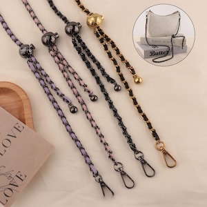 Puede incluir: Primer plano de varias correas de cadena para bolsos en varios colores. Las correas incluyen cadenas negras, doradas y moradas con herrajes metálicos a juego. Las cadenas se muestran sobre una superficie de color claro, con un pequeño bolso en el fondo.