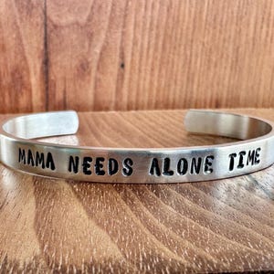 May include: A silver cuff bracelet with the stamped text "MAMA NEEDS ALONE TIME". The bracelet is made of a shiny metal and is displayed on a wooden surface.