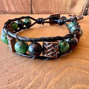 May include: A wrap bracelet featuring dark green and black beads, copper-colored square accents, and black leather straps. The bracelet is secured with a copper clasp and a leather tie closure. The beads have a marbled appearance.